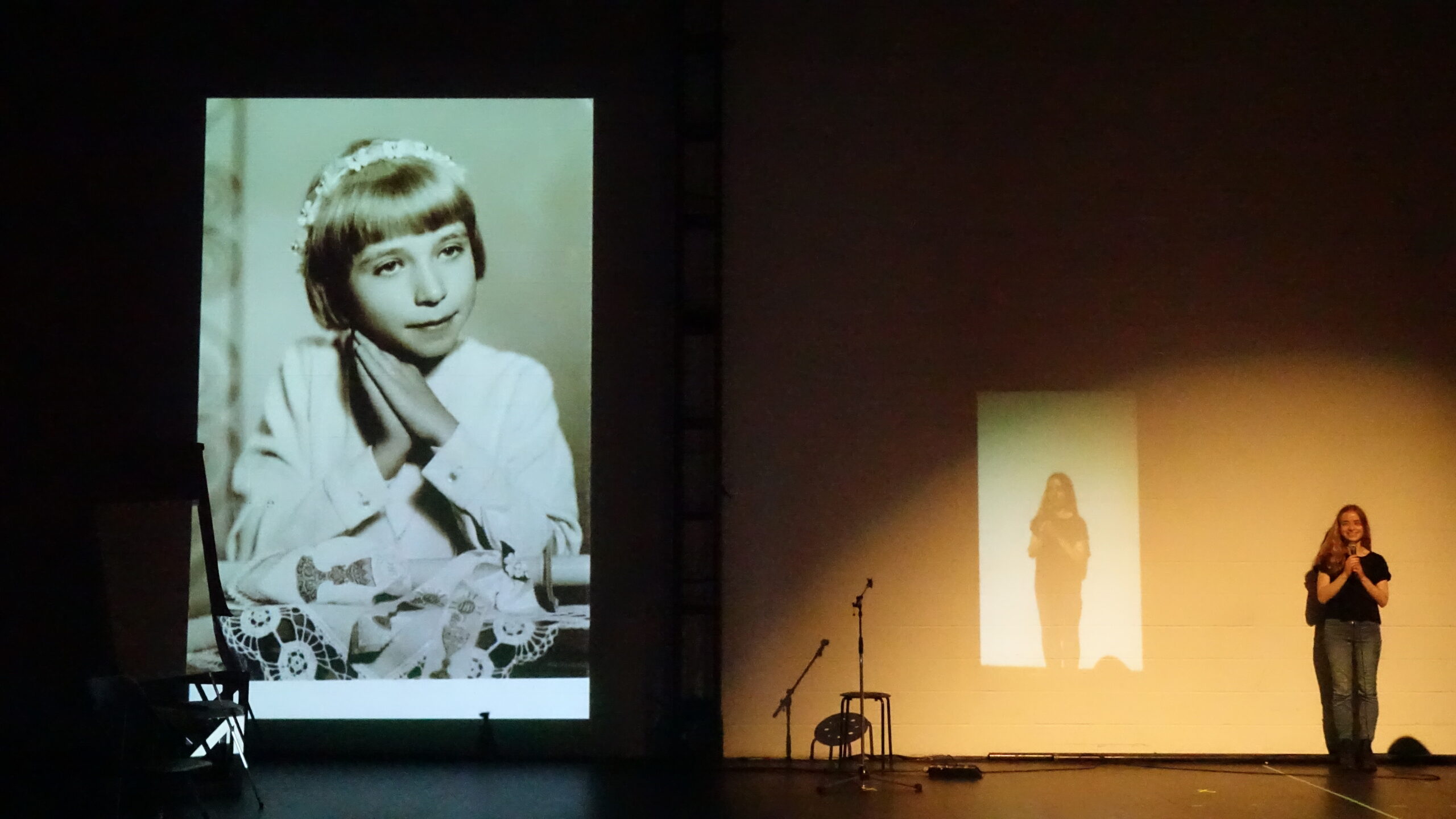 performer speaking into a microphone in front of a black-and-white projection of a young girl.