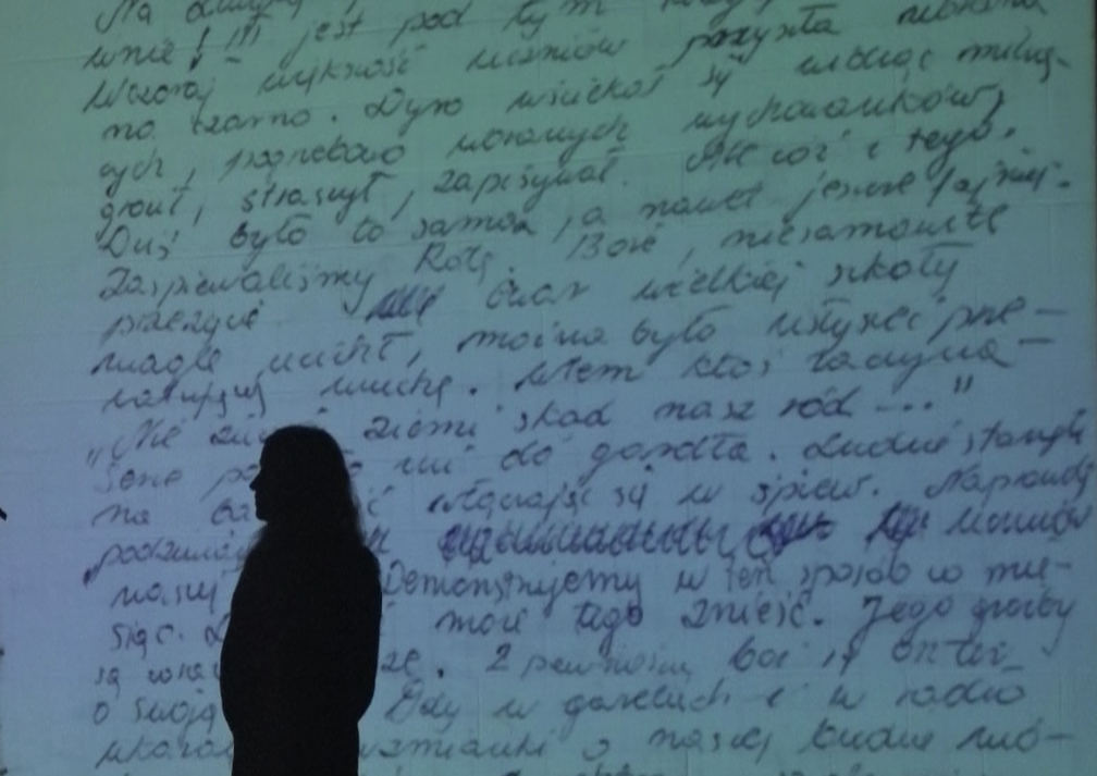 performer silhouetted against a projection of hand-written Polish text.