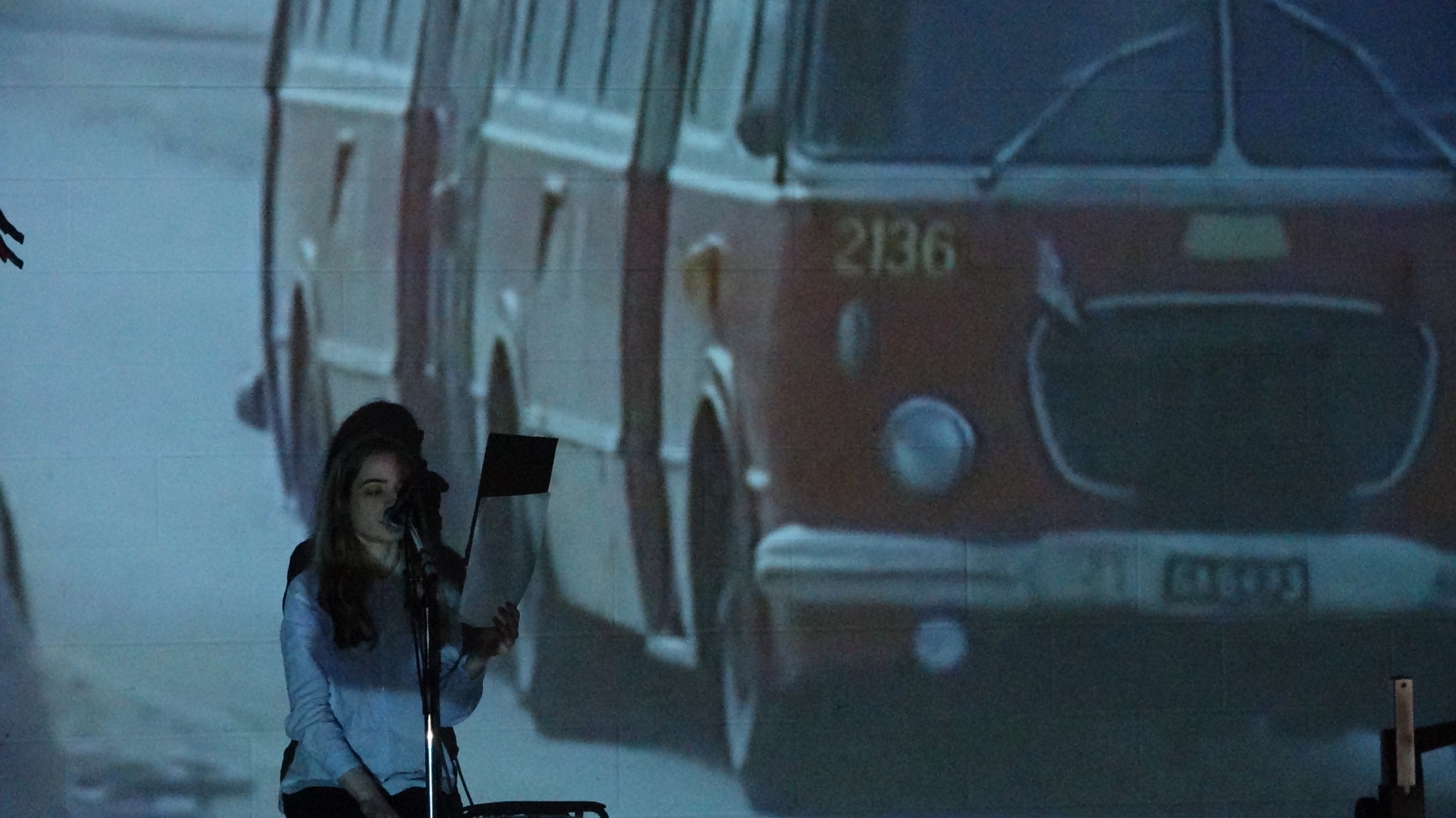 performer reading a script in front of a projection of a red, vintage bus.